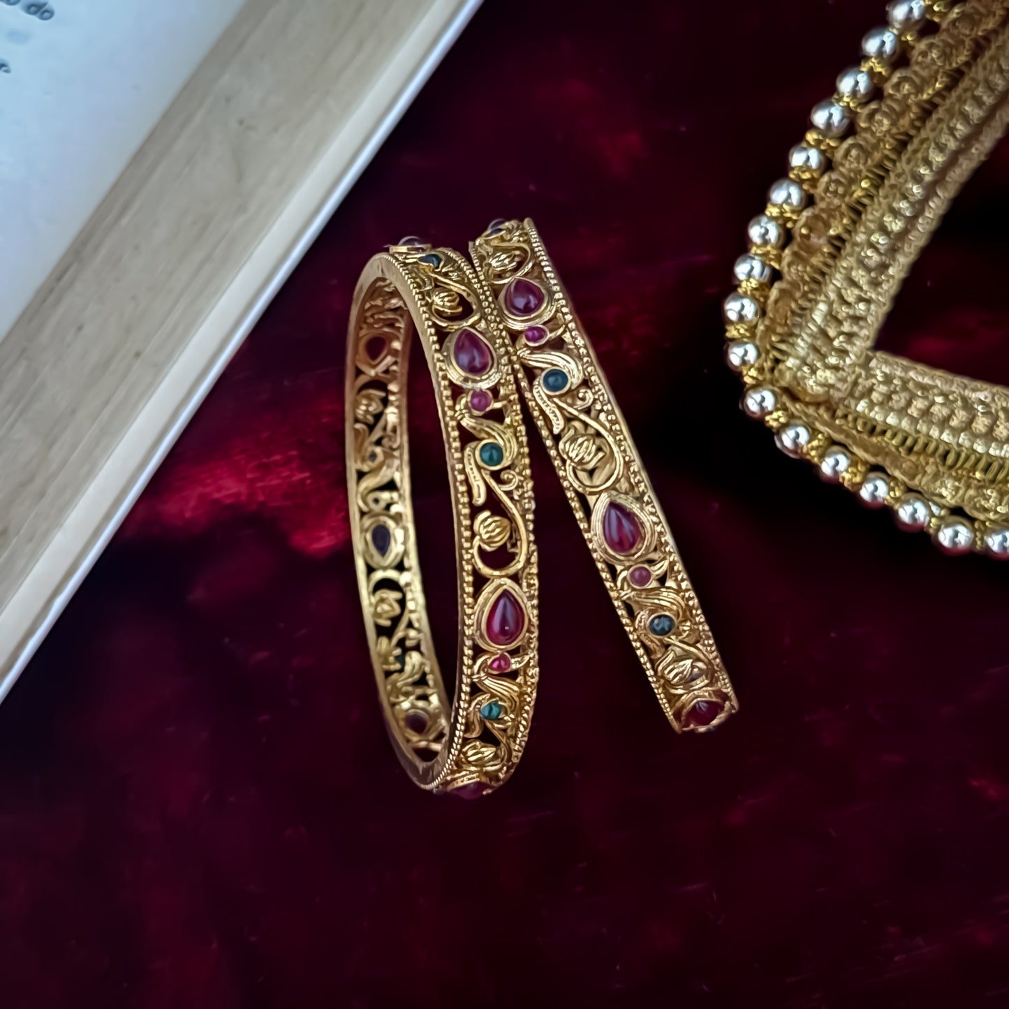 Pair of Madhavi Lakshmi Temple Gold Bangles featuring pink and green stone work and intricate antique gold filigree carving on red velvet background.