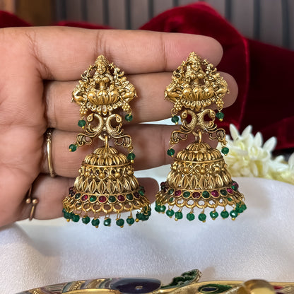 Aadya Kemp Stone premium antique gold temple earrings featuring goddess lakshmi studs twin peacock links with red and green stones and an engraved dome jhumka cup with a dense white seed pearl fringe