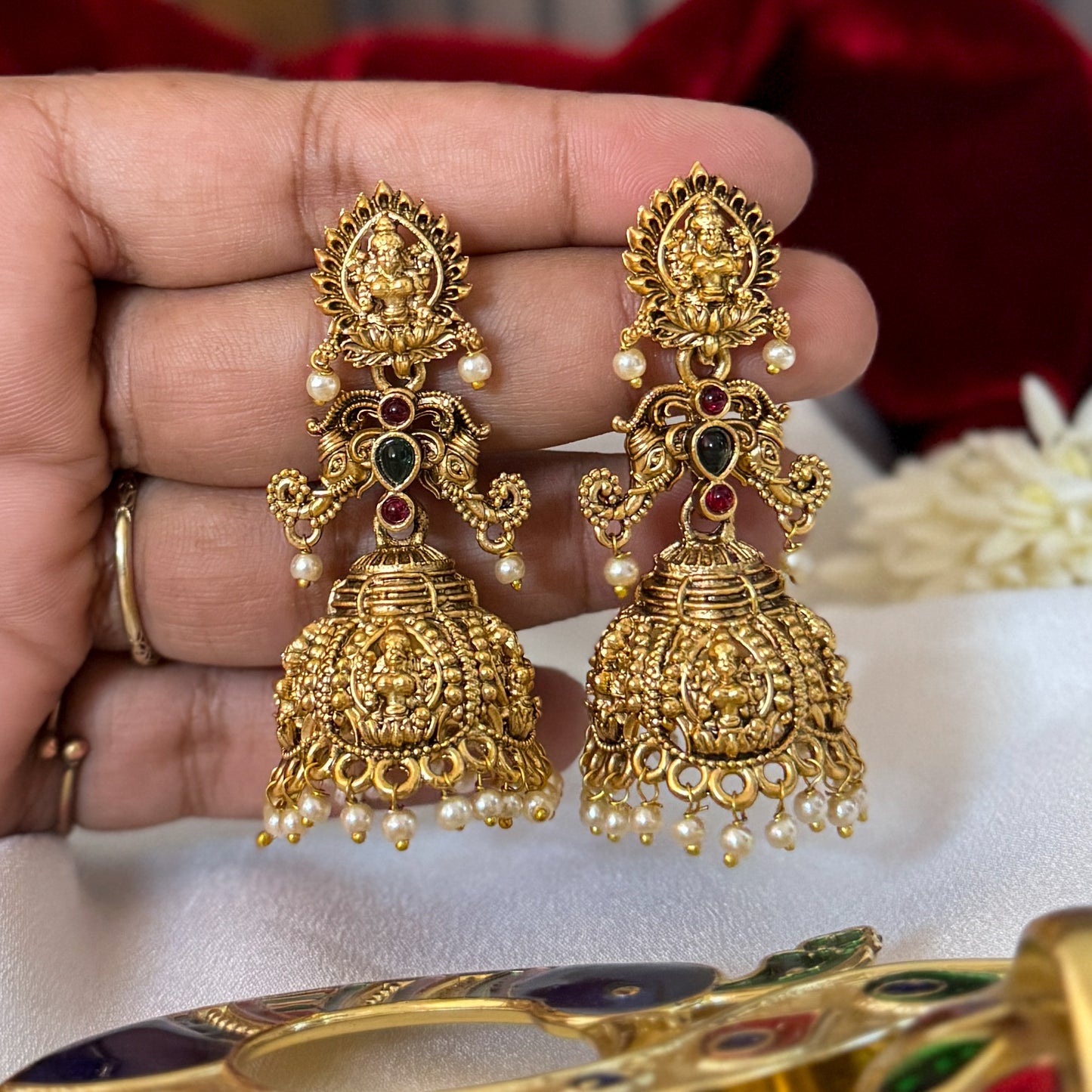 Tapasya Kemp Stone premium antique gold temple earrings featuring leaf shaped goddess lakshmi studs twin peacock links with red and green stones and an engraved dome jhumka cup with a dense white seed pearl fringe