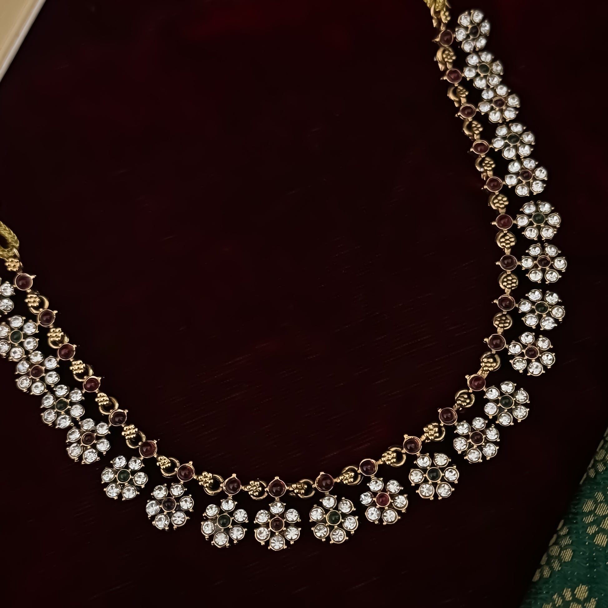 Niharika premium antique gold temple choker necklace set featuring a continuous linked row of clear crystal flower rosettes with alternating pink and green stone centers alongside matching dual flower stud earrings