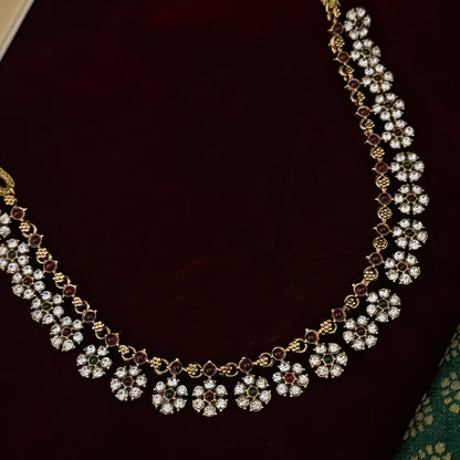 Niharika premium antique gold temple choker necklace set featuring a continuous linked row of clear crystal flower rosettes with alternating pink and green stone centers alongside matching dual flower stud earrings