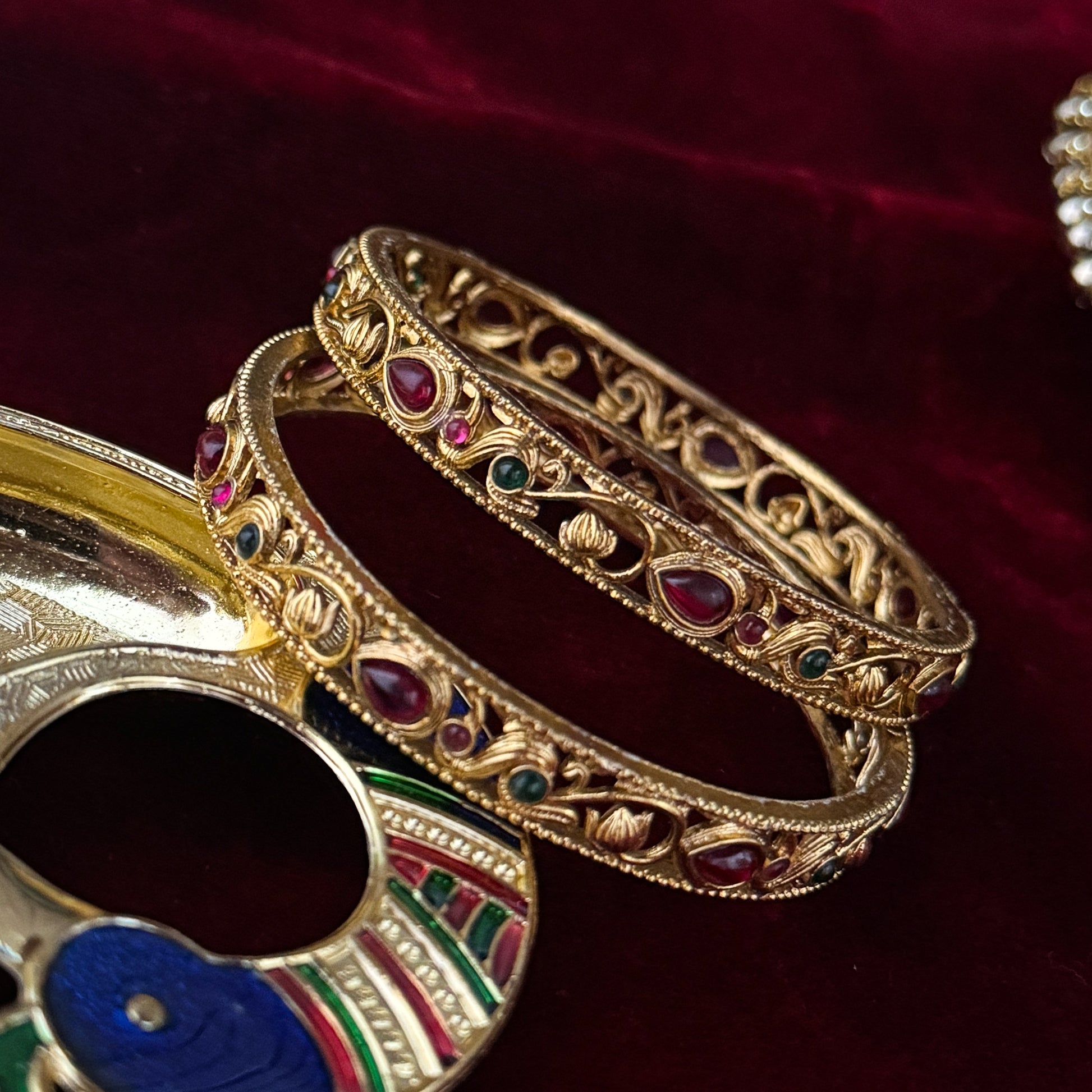 Pair of Madhavi Lakshmi Temple Gold Bangles featuring pink and green stone work and intricate antique gold filigree carving on red velvet background.