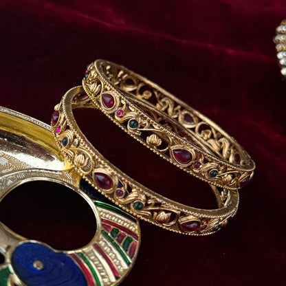 Pair of Madhavi Lakshmi Temple Gold Bangles featuring pink and green stone work and intricate antique gold filigree carving on red velvet background.