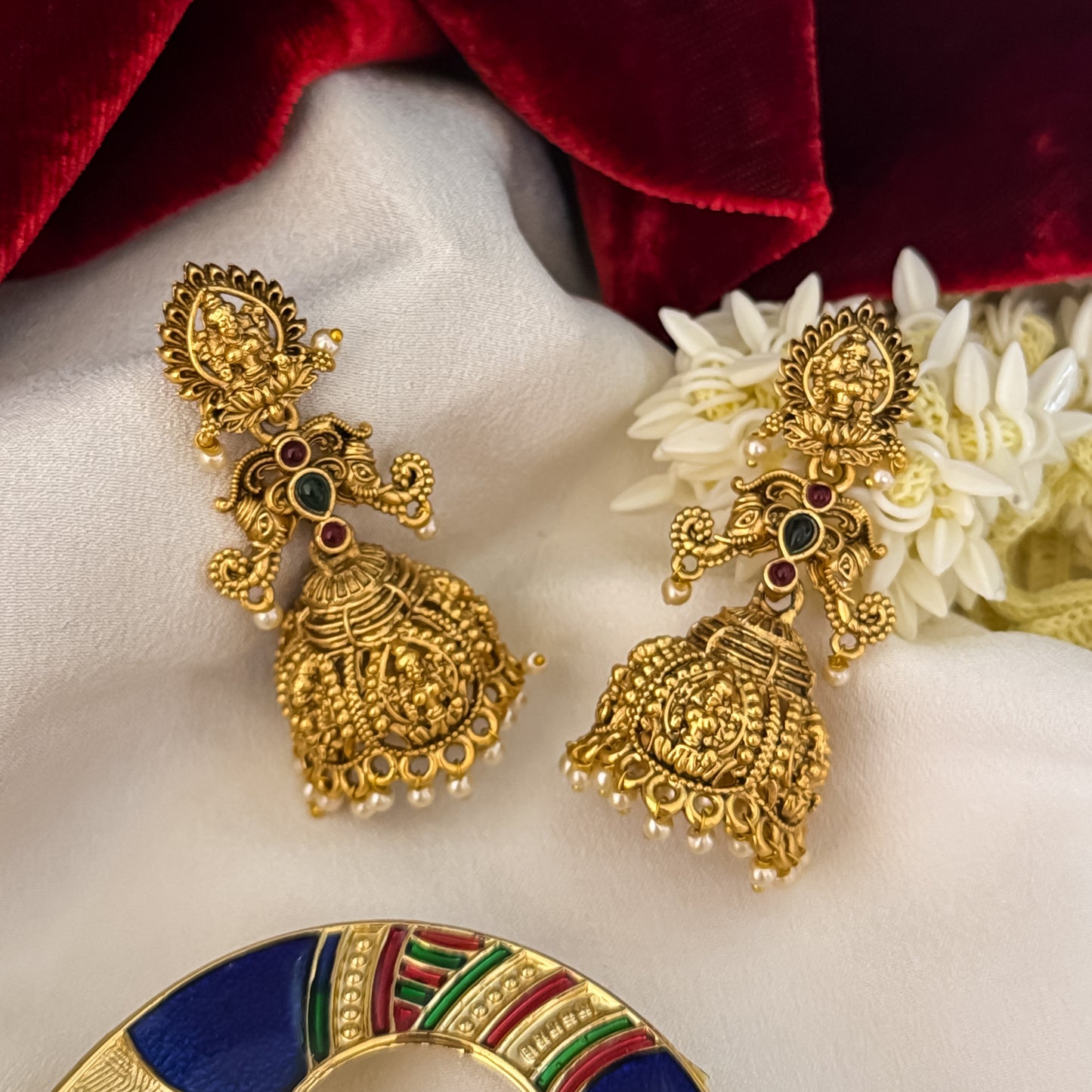 Tapasya Kemp Stone premium antique gold temple earrings featuring leaf shaped goddess lakshmi studs twin peacock links with red and green stones and an engraved dome jhumka cup with a dense white seed pearl fringe