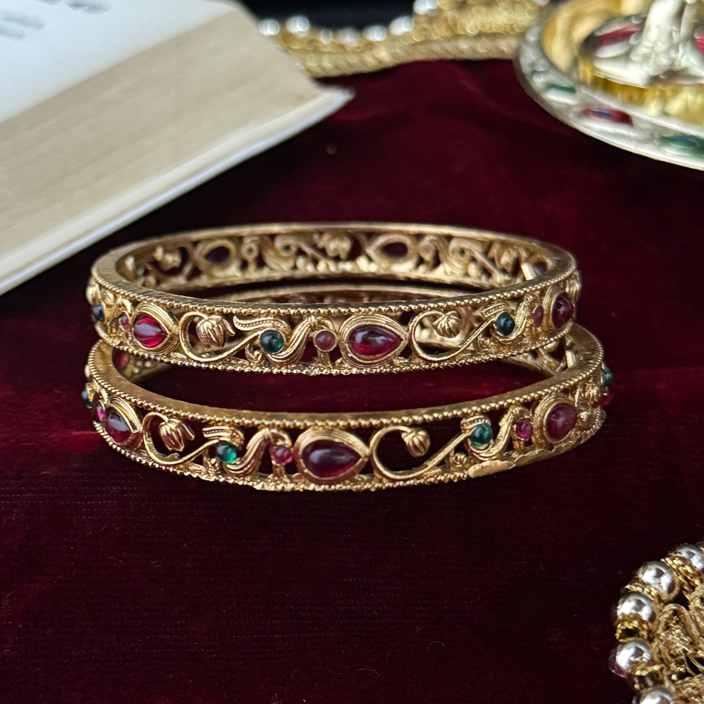 Pair of Madhavi Lakshmi Temple Gold Bangles featuring pink and green stone work and intricate antique gold filigree carving on red velvet background.