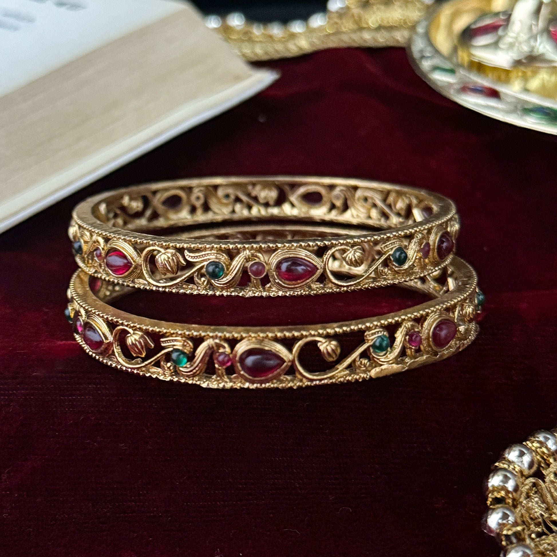 Pair of Madhavi Lakshmi Temple Gold Bangles featuring pink and green stone work and intricate antique gold filigree carving on red velvet background.
