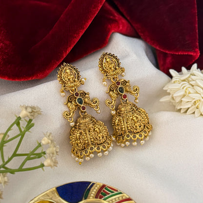 Tapasya Kemp Stone premium antique gold temple earrings featuring leaf shaped goddess lakshmi studs twin peacock links with red and green stones and an engraved dome jhumka cup with a dense white seed pearl fringe