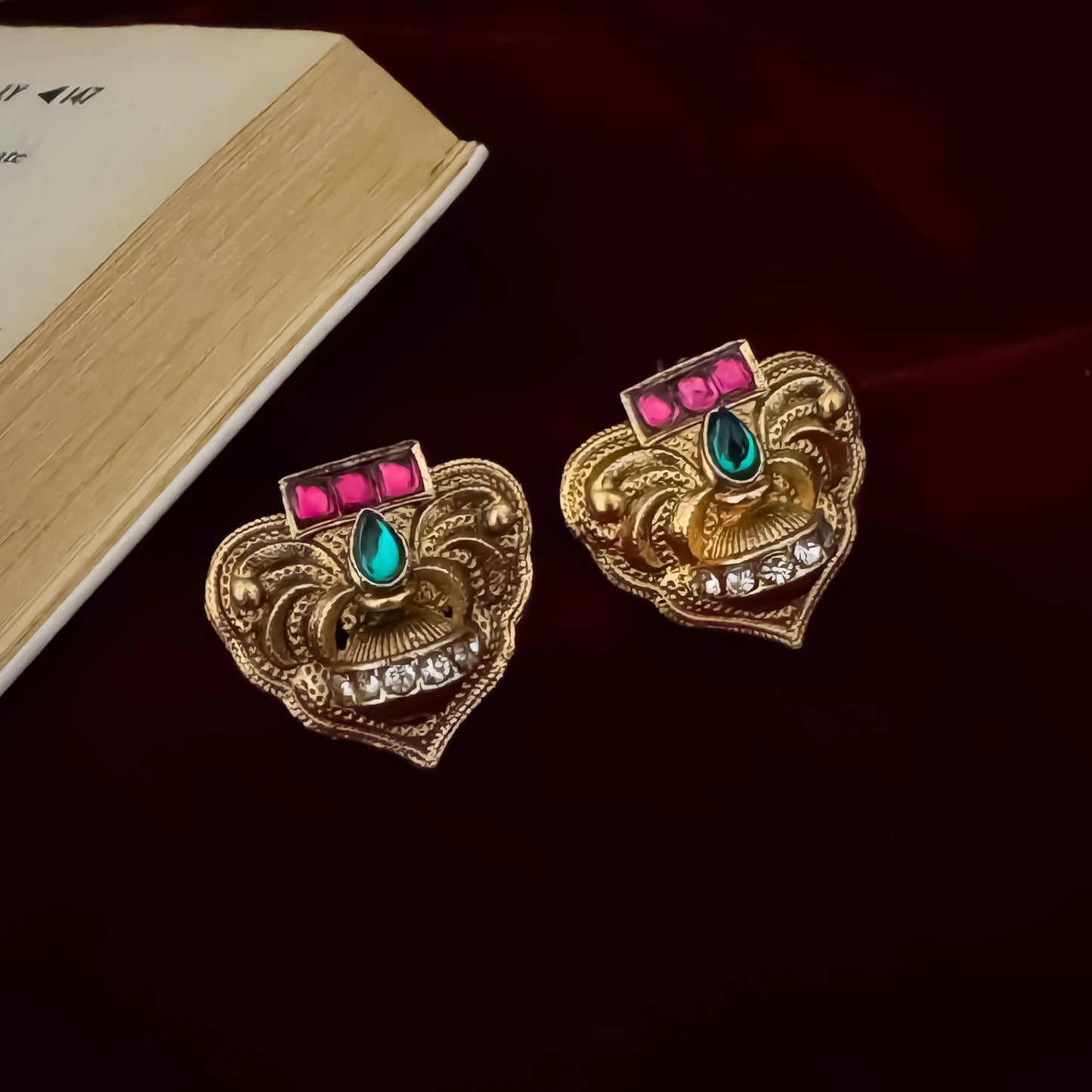 Traditional South Indian gold wedding necklace and earring set displayed flat lay over a dark red velvet background next to an open book and green silk fabric