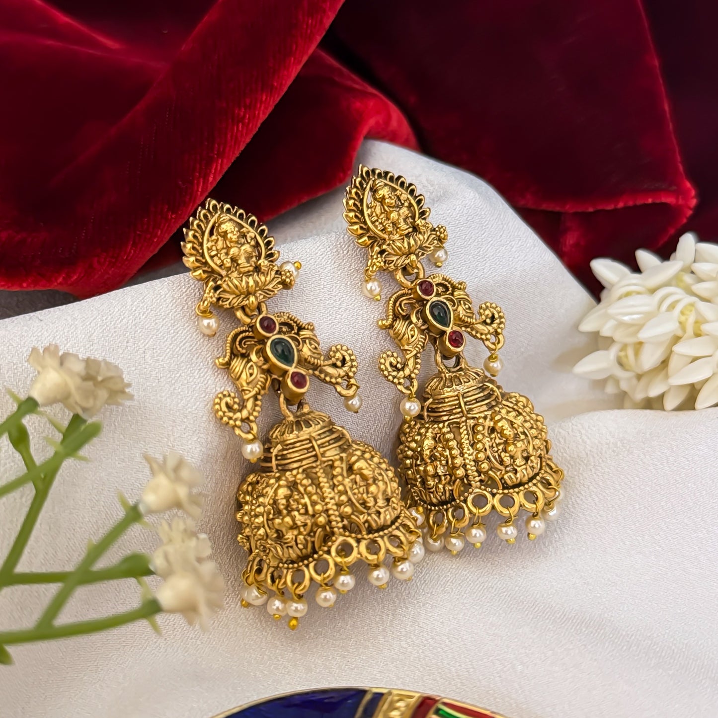 Tapasya Kemp Stone premium antique gold temple earrings featuring leaf shaped goddess lakshmi studs twin peacock links with red and green stones and an engraved dome jhumka cup with a dense white seed pearl fringe