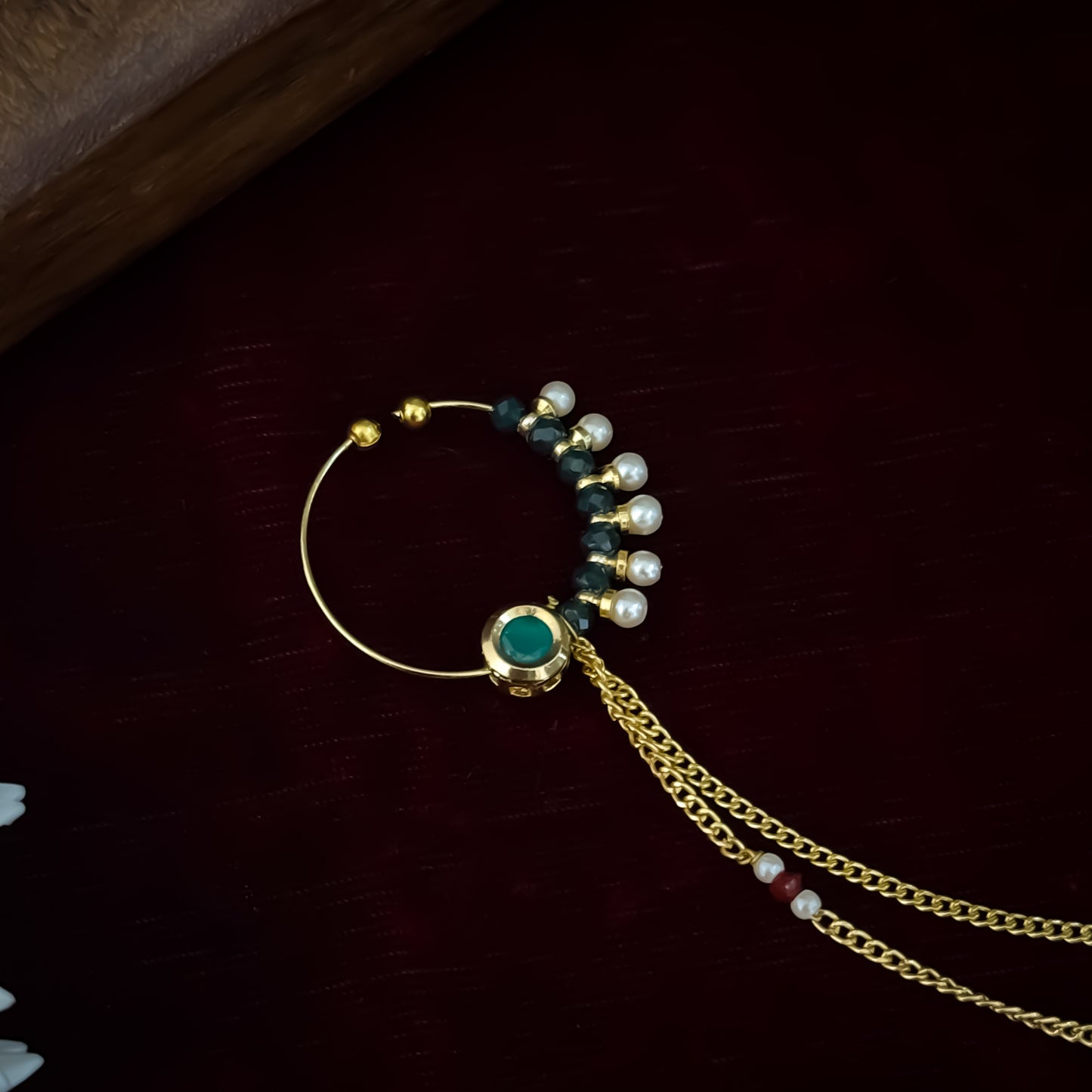 Traditional Indian wedding gold and ruby pink stone statement non pierced press on bridal nose ring nath with cascading sahara hair links displayed flat lay resting on a rich dark red maroon fabric backdrop