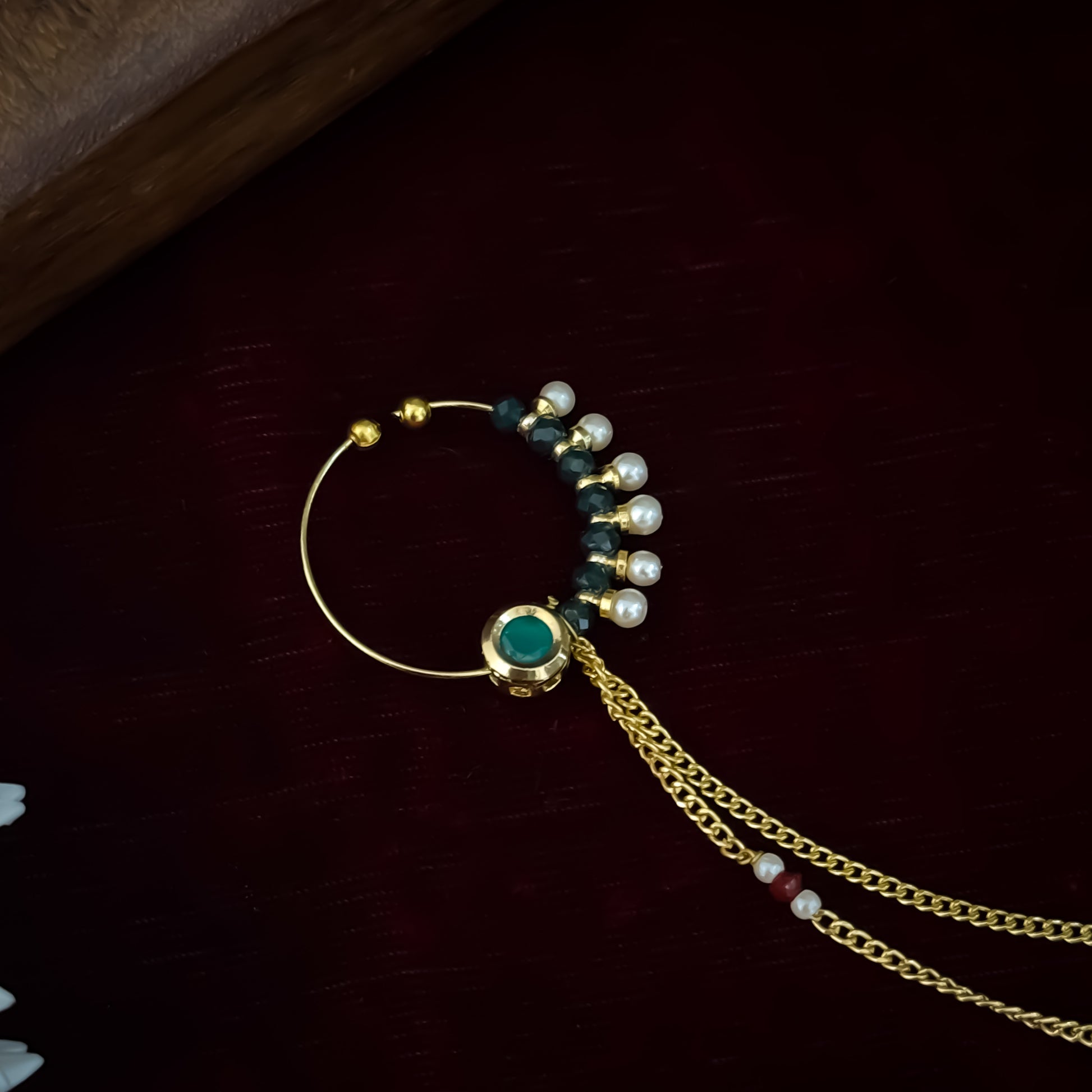 Traditional Indian wedding gold and ruby pink stone statement non pierced press on bridal nose ring nath with cascading sahara hair links displayed flat lay resting on a rich dark red maroon fabric backdrop