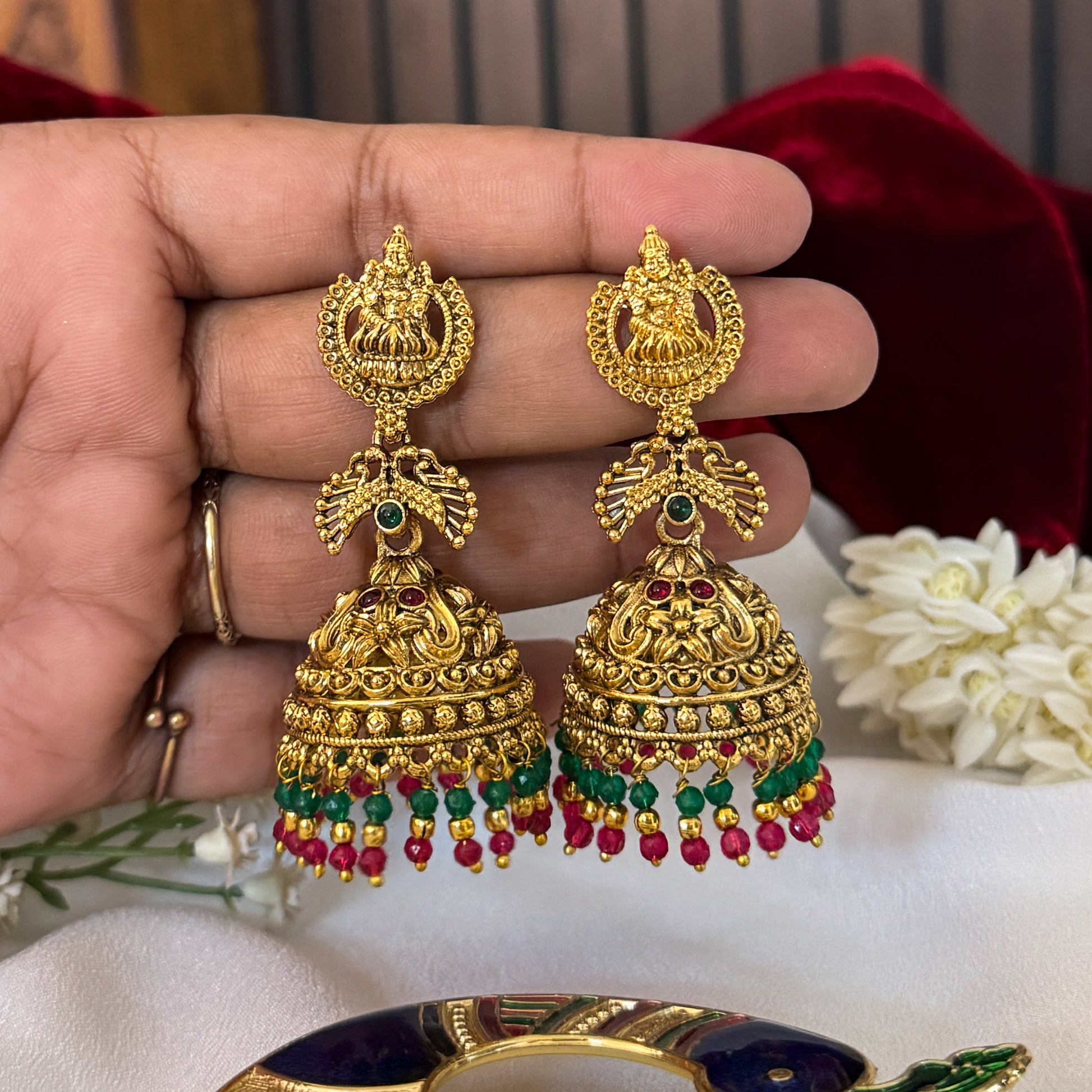 Vatsala Kemp Stone premium antique gold temple earrings featuring goddess lakshmi studs twin peacock links with red and green stones and an engraved dome jhumka cup with a dense white seed pearl fringe