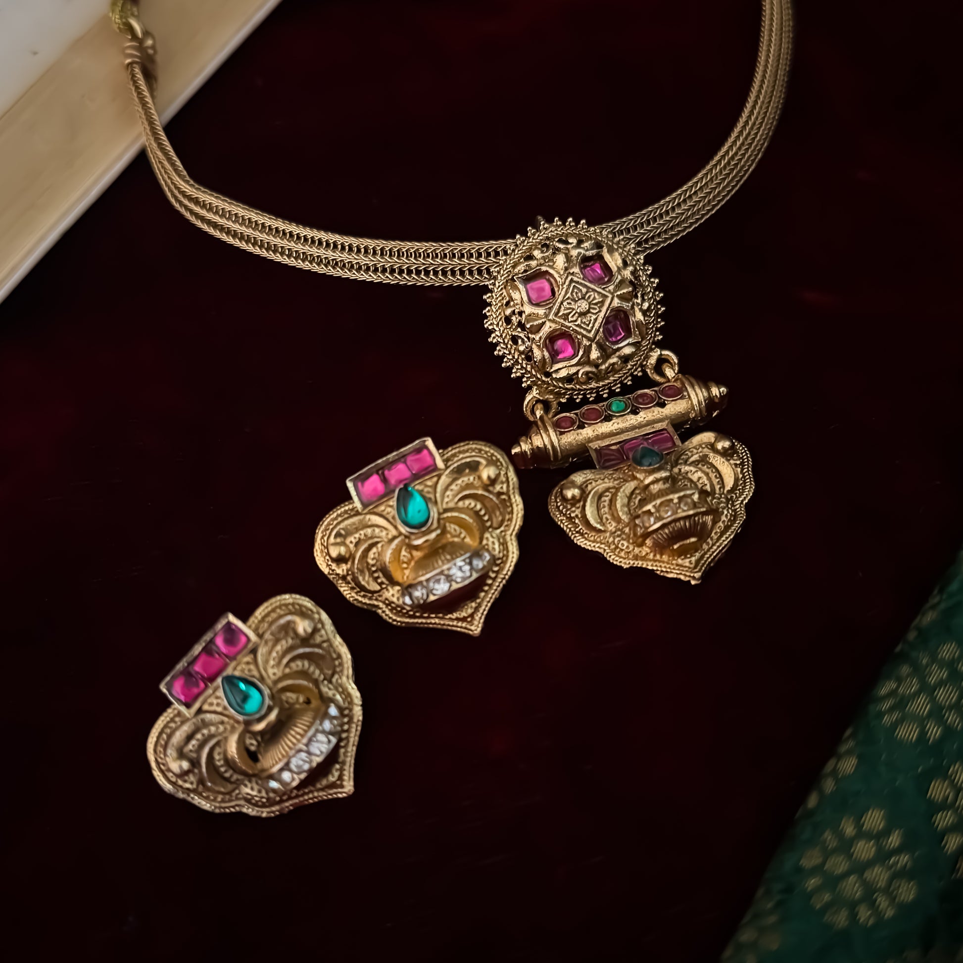 Traditional South Indian gold wedding necklace and earring set displayed flat lay over a dark red velvet background next to an open book and green silk fabric